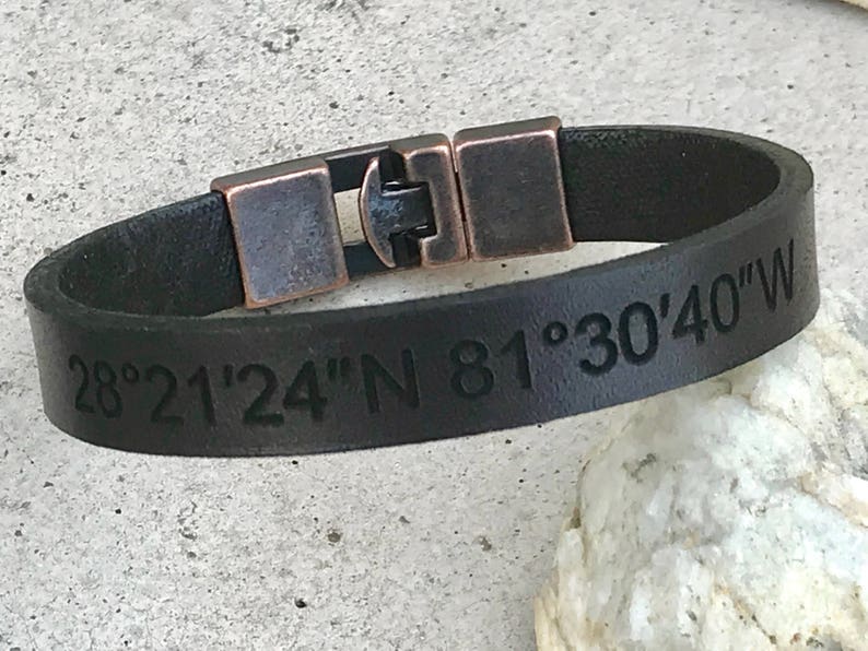 May include: Black leather bracelet with a copper buckle and engraved coordinates "28&deg;21'24"N 81&deg;30'40"W".