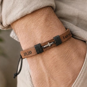 May include: A tan leather bracelet featuring a small silver-toned cross charm centered between dark grey cord wrappings. The leather band is engraved with the text 28:20 and I am wit, and includes a black adjustable cord closure.