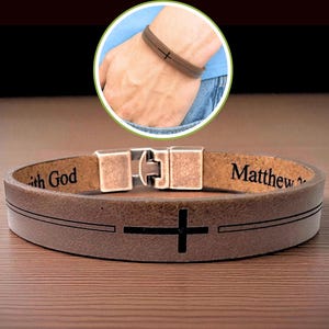 May include: A brown leather bracelet with a black cross design and the words "with God" and "Matthew" engraved on it. The bracelet has a metal clasp and is shown on a wooden surface.