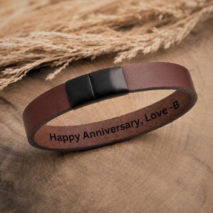 May include: A brown leather bracelet featuring a smooth matte black metal clasp. The interior surface of the leather band is engraved with the text "Happy Anniversary, Love -B" in a clear, dark, sans-serif font, visible along the inner side.