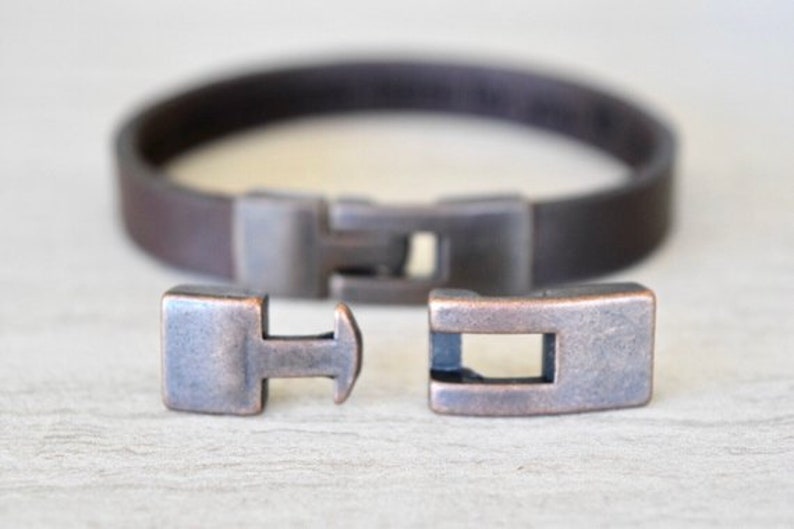 May include: A brown leather bracelet with a copper-toned metal clasp. The clasp is made of two pieces that fit together.