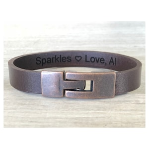 May include: A brown leather bracelet with a copper clasp. The bracelet is engraved with the text "Sparkles Love, Al".