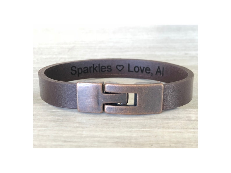 May include: A brown leather bracelet with a copper clasp. The bracelet is engraved with the text "Sparkles Love, Al".