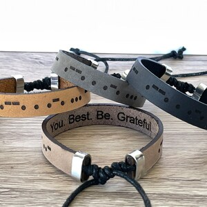 May include: Four leather bracelets with adjustable closures. Each bracelet features a different color: brown, gray, black, and tan. The tan bracelet has the text "You. Best. Be. Grateful" engraved on it. All bracelets have braille dots engraved on them.