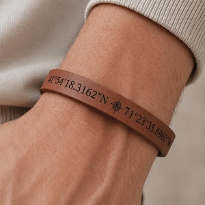 May include: A smooth, tan leather bracelet featuring black engraved text that reads 41°54'18.3162"N 71°23'35.6960"W, with a small compass rose icon positioned between the two sets of coordinates, encircling the wrist in a simple, flat band design.