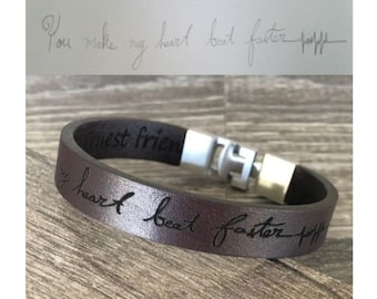 May include: A brown leather bracelet with the engraved text "You make my heart beat faster" and a silver clasp. The bracelet is on a wooden surface. Above the bracelet is a handwritten note with the same text.