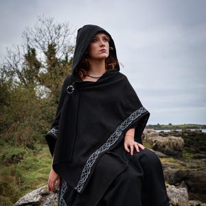 Könnte beinhalten: Schwarzer Kapuzenponcho mit silberner Schließe und Besatz. Der Poncho wird über einer schwarzen Hose getragen. Der Besatz weist ein weißes, geometrisches Design auf. Die Person sitzt auf einem Felsen, mit bewölktem Himmel im Hintergrund.