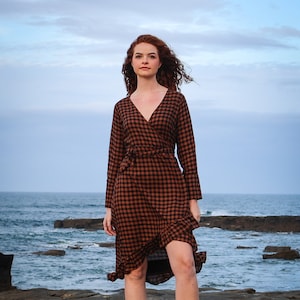 Könnte beinhalten: Eine Frau in einem braunen und schwarzen karierten Wickelkleid mit gerafftem Saum und braunen Lederstiefeln. Sie steht auf einem felsigen Strand mit dem Meer im Hintergrund.