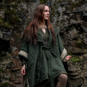 May include: A woman wearing a long, dark green hooded cloak with a brown belt and intricate embroidery. She is standing in front of a stone wall with moss growing on it. She is wearing brown leather boots.