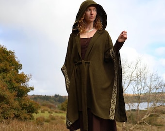 May include: A woman wearing a dark green hooded cloak with gold trim, standing on a mossy rock. The cloak is tied at the waist with a matching belt. The woman is wearing a brown dress and boots. The background features a landscape with trees and a lake.
