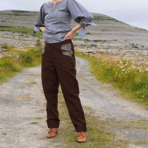 Op de afbeelding: Een vrouw draagt een lichtblauwe blouse met een kanten halslijn en een bruine broek met geruite zakken. Ze staat op een onverhard pad met een grasveld en een rotsachtige heuvel op de achtergrond.
