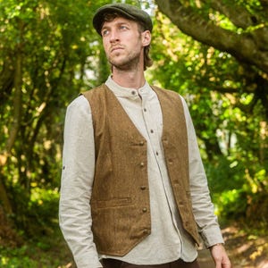 Könnte beinhalten: Eine braune Tweedweste mit V-Ausschnitt und Knopfverschluss, getragen über einem hellen Button-Down-Hemd. Die Person trägt auch eine grüne Mütze. Das Outfit ist für Outdoor-Aktivitäten geeignet.