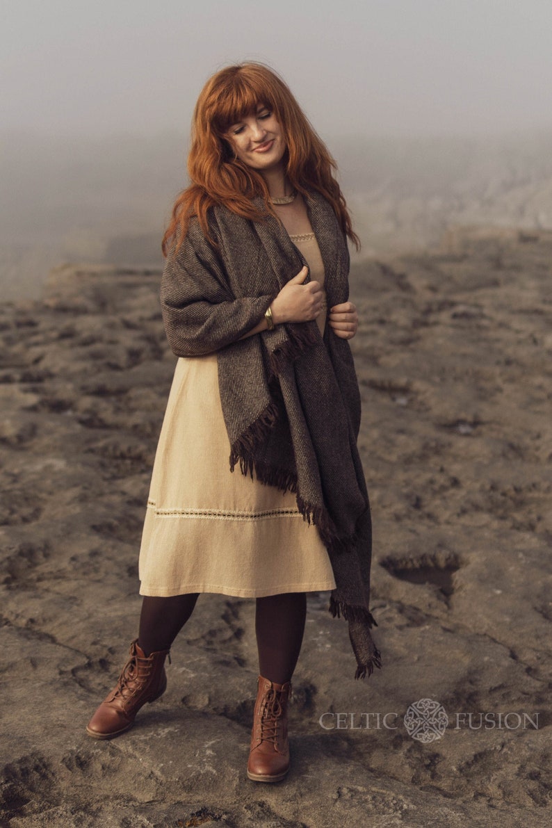 Pu&ograve; includere: Un abito beige al ginocchio con un dettaglio in pizzo orizzontale, abbinato a uno scialle intrecciato marrone scuro con bordi sfrangiati, collant marrone scuro e stivaletti stringati in pelle marrone. Il testo Celtic Fusion &egrave; visibile nell'angolo.