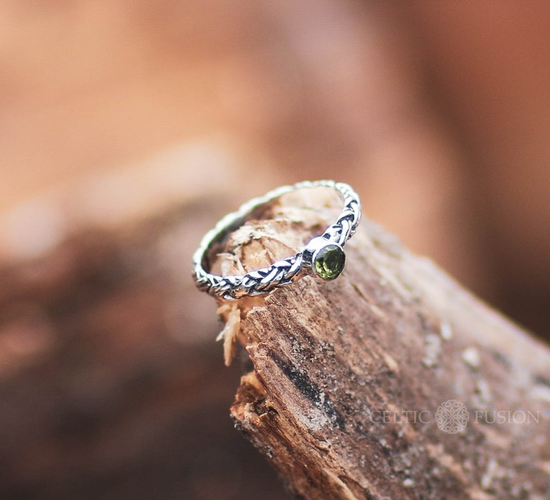 May include: A silver ring with a braided design and a green gemstone. The ring is sitting on a piece of weathered wood.