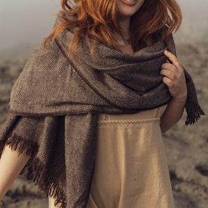 Pu&ograve; includere: Uno scialle con motivo a spina di pesce marrone scuro e beige con bordi sfrangiati drappeggiato su un abito beige senza maniche. L'angolo in basso a destra presenta il testo CELTIC FUSION con un logo circolare a intreccio stampato in bianco.