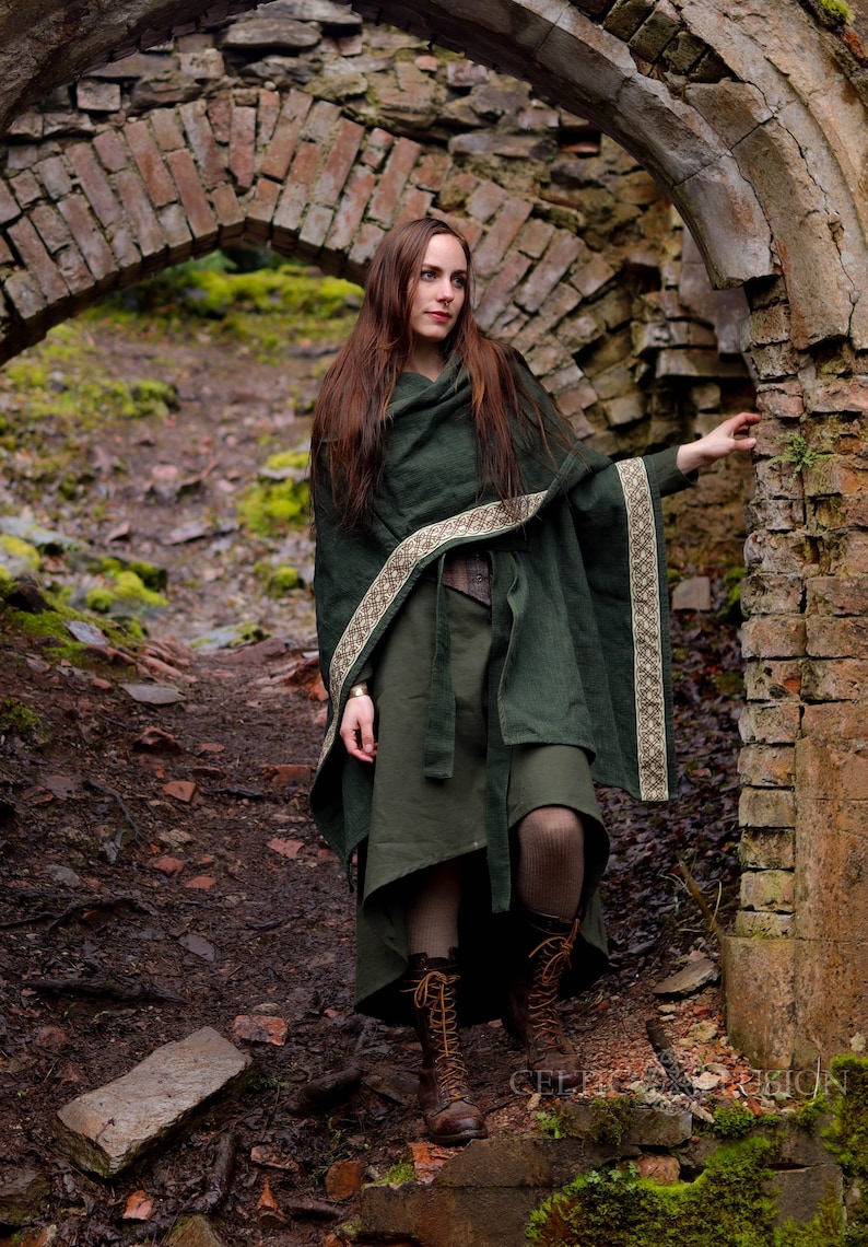 May include: A woman wearing a dark green hooded cloak with a brown and gold trim stands in a stone archway. The cloak is tied at the waist with a brown belt. The woman is wearing brown boots and has long brown hair.