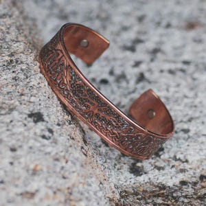 May include: A copper-colored cuff bracelet with an intricate Celtic knot design. The open-ended bracelet features a detailed pattern across its surface. The metal has a slightly aged appearance, and the bracelet is resting on a textured gray surface.