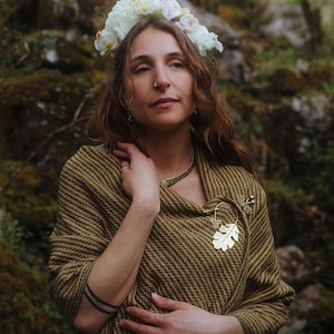 May include: A gold-toned metal brooch shaped like an oak leaf with spiral accents, pinned to a textured olive green and black woven shawl. The brooch features a delicate stem and small leaf details, securing the fabric with a simple metal pin mechanism.