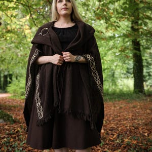 May include: A dark brown hooded cloak featuring intricate cream-colored Celtic knotwork embroidery along the front edges and a fringed hem. The garment is secured at the shoulder with a circular metal brooch and worn over a black dress and boots.