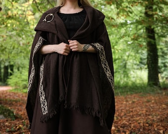 May include: A dark brown hooded cloak featuring intricate cream-colored Celtic knotwork embroidery along the front edges and a fringed hem. The garment is secured at the shoulder with a circular metal brooch and worn over a black dress and boots.