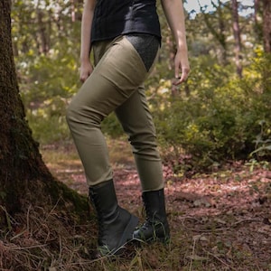 May include: Olive green pants with a dark gray panel on the upper thigh. The pants are paired with black, lace-up boots. The person is standing in a forest setting with trees and foliage in the background. The pants have a slim fit.