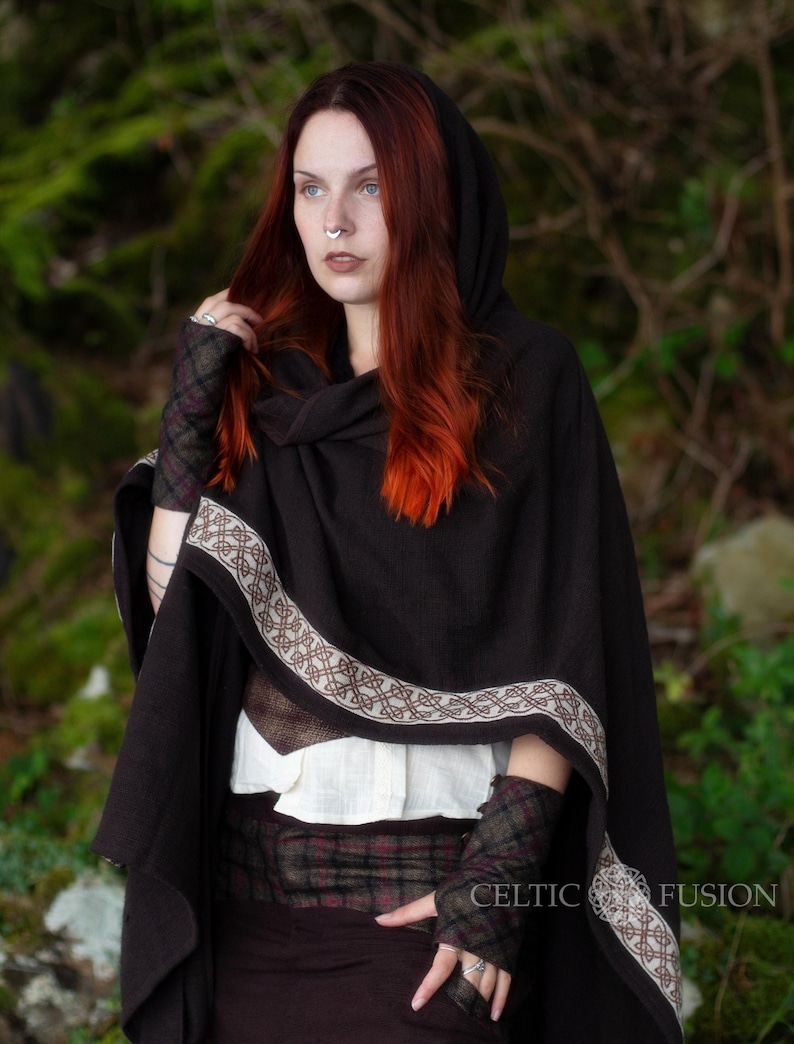 May include: A brown hooded cloak with a Celtic knot trim and matching fingerless gloves. The cloak is worn over a white shirt and a plaid skirt. The model is wearing a silver nose ring.