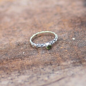 May include: A silver ring with a braided band and a green gemstone. The ring is on a wooden surface.