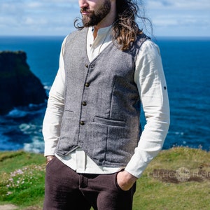 Op de afbeelding: Een grijze tweed vest met vier knopen en twee voorzakken. Het wordt gedragen over een crèmekleurig shirt met lange mouwen en een bruine broek. De achtergrond toont een kustlandschap met blauw water en groene kliffen.