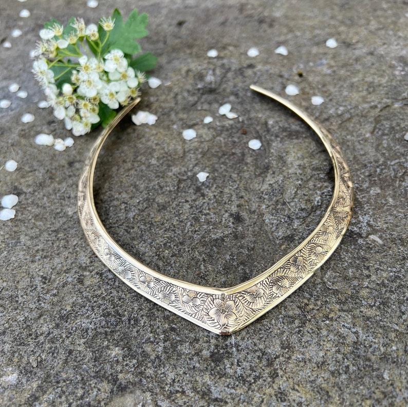 May include: A gold-toned metal choker necklace with a floral design. The necklace is on a grey textured surface.