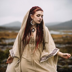 May include: A woman with long red hair wearing a beige linen dress with a lace-up bodice and a long, flowing beige shawl with black embroidery. She is wearing a silver crescent moon necklace and earrings.