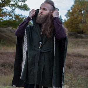 May include: A hooded green cloak with white and brown trim, tied at the waist with a brown belt. The cloak has a Celtic knot design on the trim. The person wearing the cloak has long brown hair and a beard.