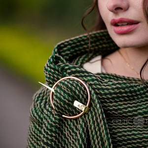 May include: A green and beige woven shawl with a gold metal ring and pin closure. The shawl is worn by a person with long brown hair. The text "CELTIC FUSION" is visible in the lower right corner of the image.