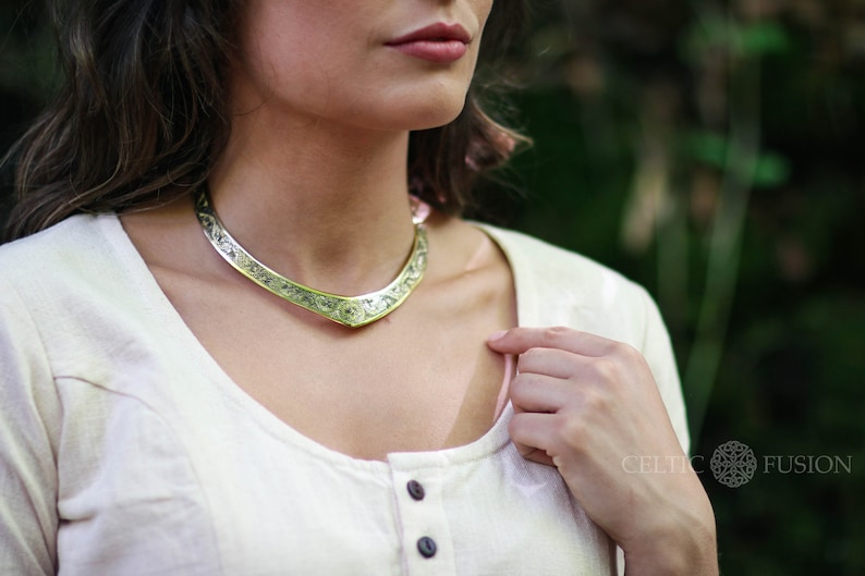 May include: A silver Celtic knot choker necklace with a detailed design. The necklace is worn by a person wearing a white shirt.