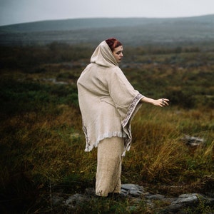 May include: A woman wearing a hooded, beige, crocheted poncho with a fringe hem. The poncho has a dark brown embroidered design. She is standing in a field of tall grass.