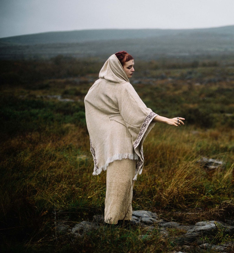 May include: A woman wearing a hooded, beige, crocheted poncho with a fringe hem. The poncho has a dark brown embroidered design. She is standing in a field of tall grass.