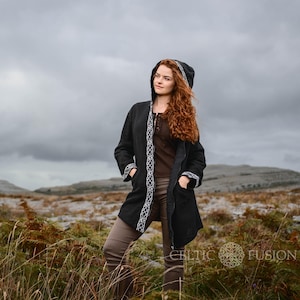 Könnte beinhalten: Eine schwarze Kapuzenjacke mit weißen keltischen Knoten Stickereien. Die Jacke hat ein langes, fließendes Design und wird von einer Person mit langen, roten Haaren getragen. Die Person steht auf einem Feld mit einem bewölkten Himmel im Hintergrund.