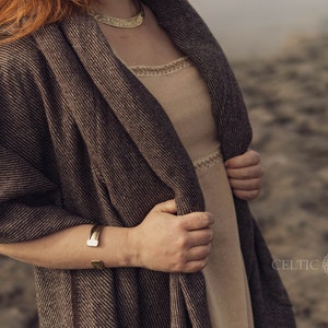 Pu&ograve; includere: Uno scialle testurizzato marrone e nero drappeggiato su un abito beige con un bordo color crema sullo scollo e in vita. L'insieme include una collana girocollo in metallo dorato e un bracciale a fascia in metallo dorato abbinato al polso.
