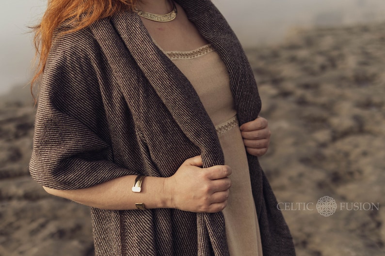 Pu&ograve; includere: Uno scialle testurizzato marrone e nero drappeggiato su un abito beige con un bordo color crema sullo scollo e in vita. L'insieme include una collana girocollo in metallo dorato e un bracciale a fascia in metallo dorato abbinato al polso.