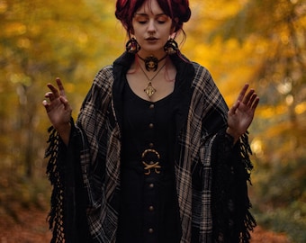 May include: A woman with red hair wearing a black dress with a plaid shawl and a gold belt. She is standing in a forest with fall foliage.