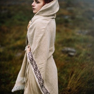 May include: A woman with red hair wearing a hooded, beige shawl with a dark brown braided trim. The shawl has a fringe along the bottom edge. The woman is standing in a field of tall grass.