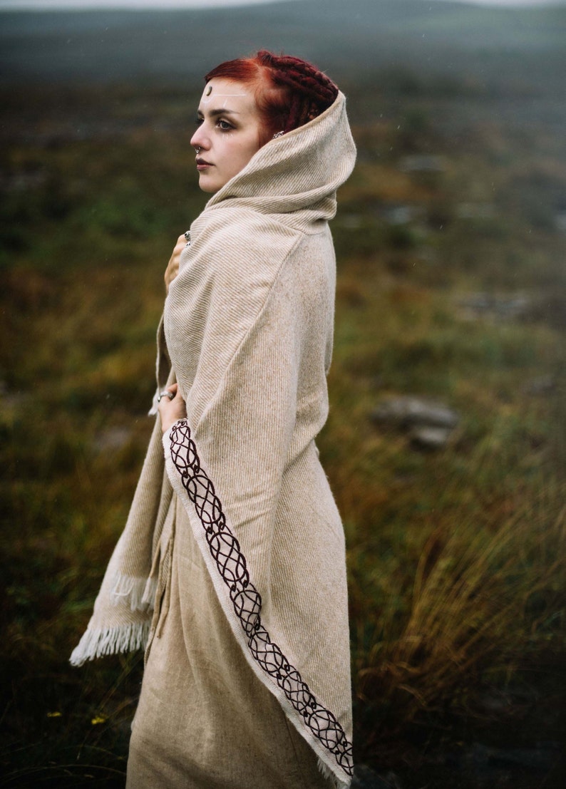 May include: A woman with red hair wearing a hooded, beige shawl with a dark brown braided trim. The shawl has a fringe along the bottom edge. The woman is standing in a field of tall grass.