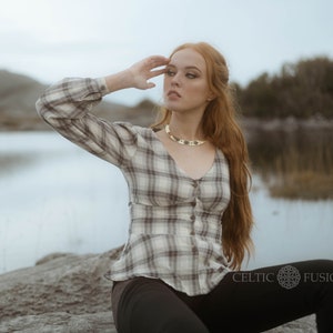 Op de afbeelding: Een vrouw draagt een wit en bruin geruit overhemd met een gouden ketting. Ze zit op een rots met een waterpartij achter haar. De tekst "Celtic Fusion" is zichtbaar in de rechterbenedenhoek van de afbeelding.