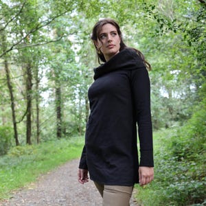 Puede incluir: Una túnica negra de manga larga con cuello alto. La túnica está hecha de un material suave y tiene un corte holgado. La persona lleva pantalones color beige y está de pie en un camino en un bosque.