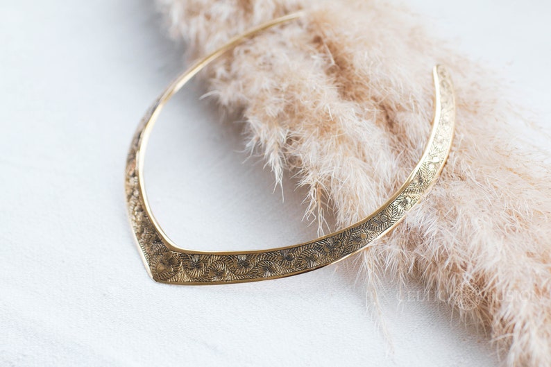 May include: A gold-toned metal necklace with an intricate floral design. The necklace is lying on a soft, fluffy surface.