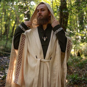 Op de afbeelding: Een crèmekleurige cape met capuchon met ingewikkelde geweven details en een bijpassende riem. De cape heeft decoratieve randen in een roodbruine tint langs de randen. De persoon draagt een donkergroen shirt en een hanger. De achtergrond is een bos.