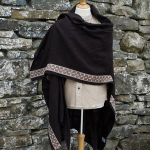 May include: A brown hooded shawl with a decorative brown and cream trim. The shawl is draped over a mannequin and is displayed against a stone wall.