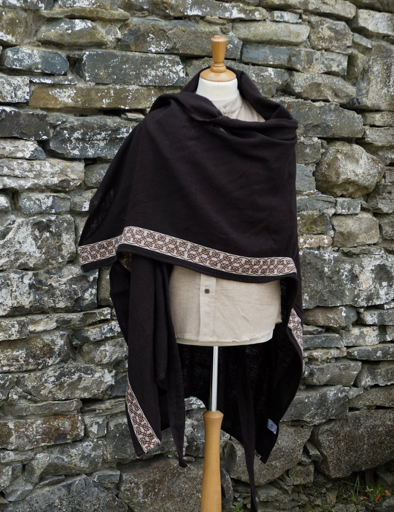 May include: A brown hooded shawl with a decorative brown and cream trim. The shawl is draped over a mannequin and is displayed against a stone wall.