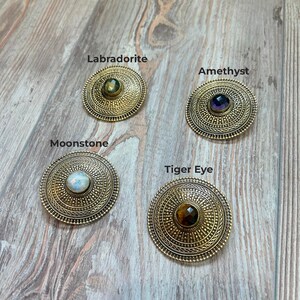 May include: Four antique gold-colored, circular brooches with different gemstones: Labradorite, Amethyst, Moonstone, and Tiger Eye. Each brooch has a detailed, textured design around the central stone, with the gemstone names printed above each brooch.