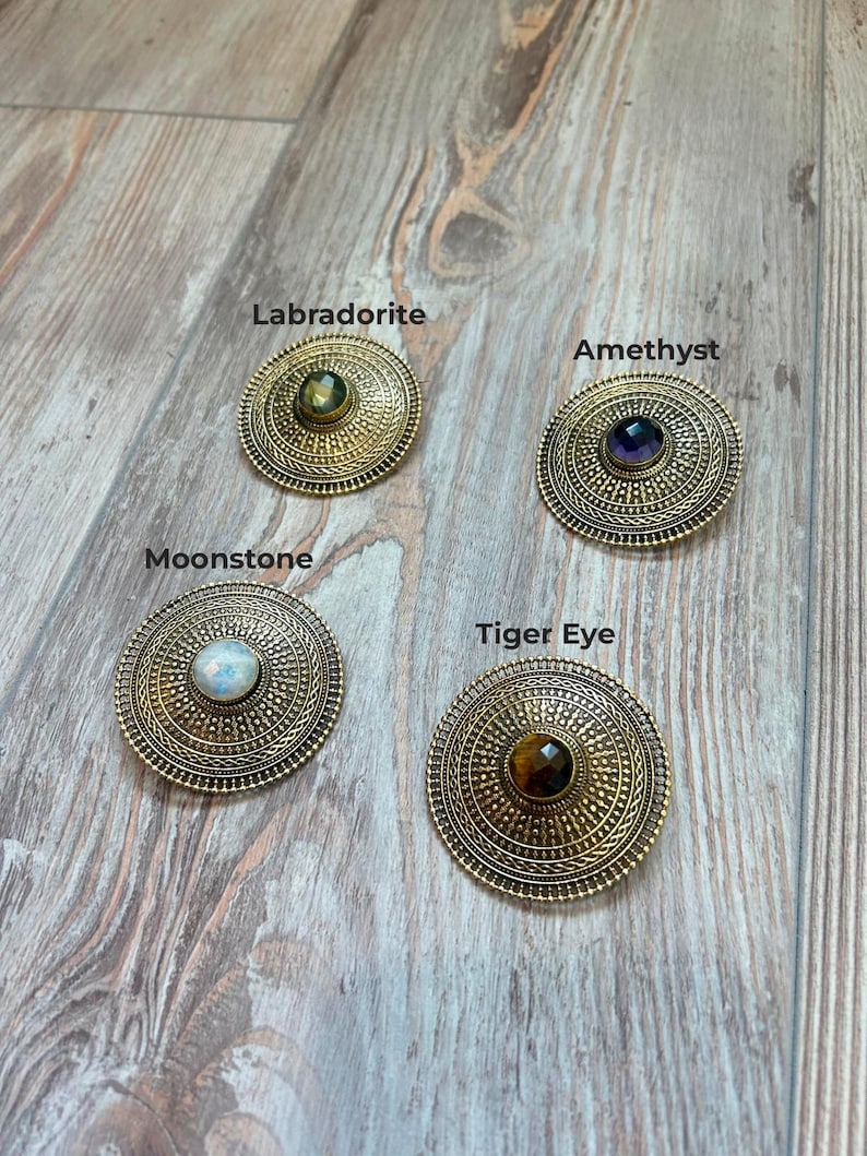 May include: Four antique gold-colored, circular brooches with different gemstones: Labradorite, Amethyst, Moonstone, and Tiger Eye. Each brooch has a detailed, textured design around the central stone, with the gemstone names printed above each brooch.