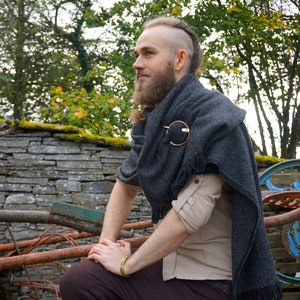 May include: A dark gray woven shawl with fringe edges, secured by a circular gold-toned metal brooch with a twisted texture. The shawl is draped over a light beige button-down shirt with rolled sleeves and dark brown trousers, paired with a gold-toned cuff.
